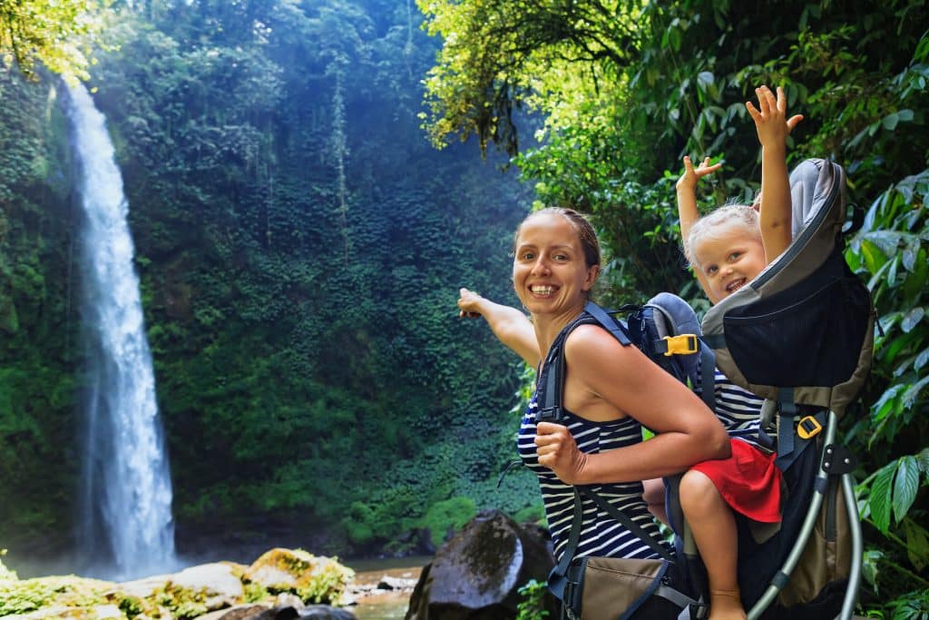 In waterfall pool young happy mother hold little traveller on back - baby girl in carrying backpack enjoying travel adventure Hiking activity with child on family summer vacation weekend nature tour