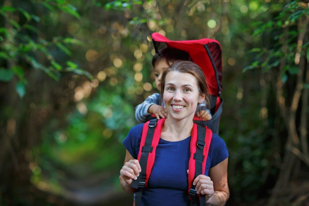 Mother with toddler child in backpack carrier is hiking in forest. Tourist is carrying a baby on his back in the natural park