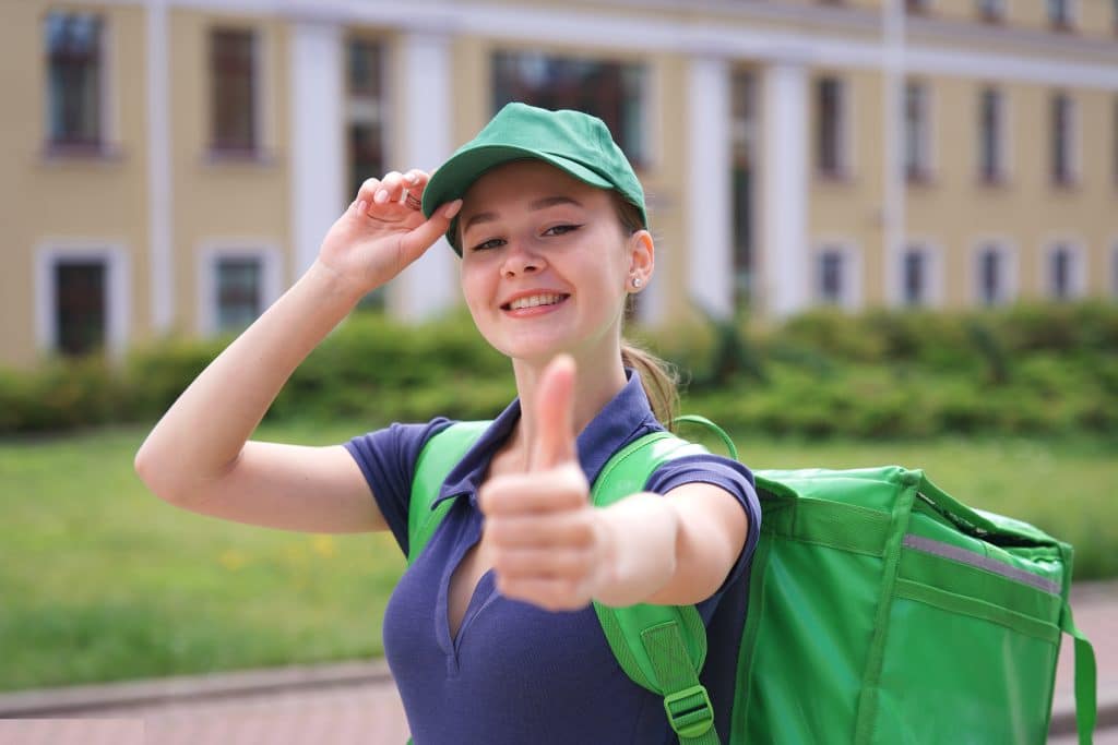 portrait of young beautiful happy courier teenager girl, food delivery woman with green thermo box for food delivering food outdoors in the yard at summer day in cap and uniform. Student job, work