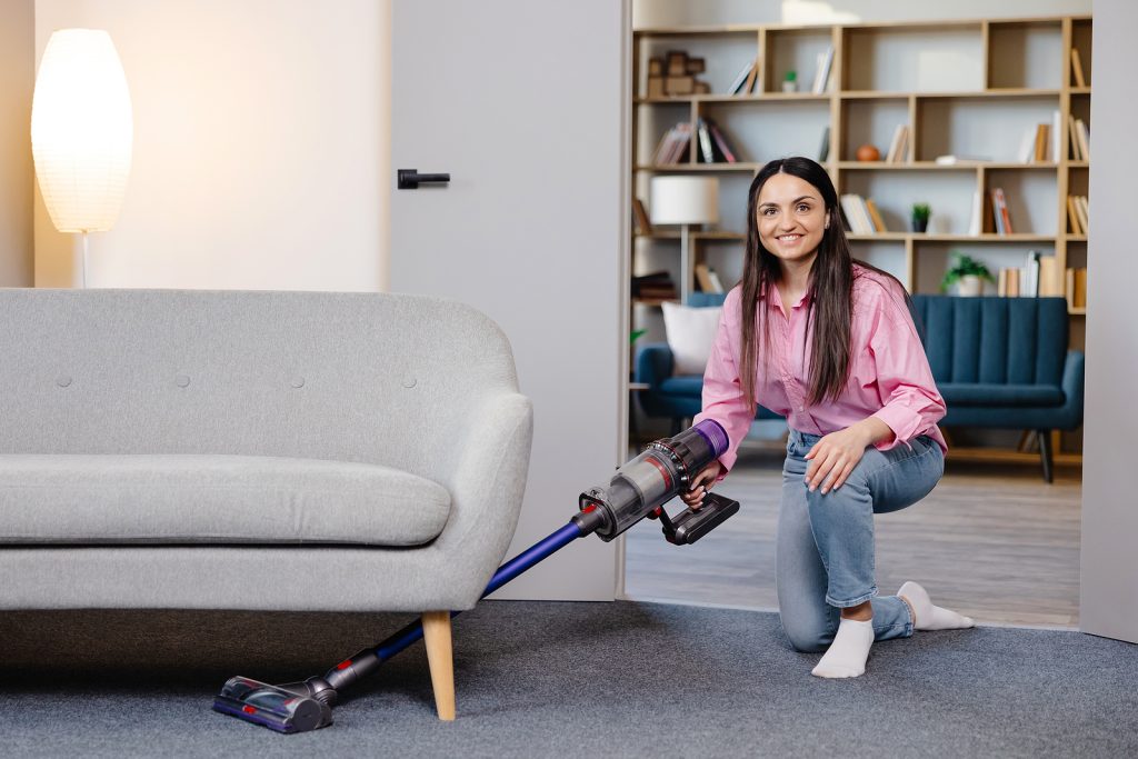 Young woman uses cordless vacuum cleaner to clean home carpet