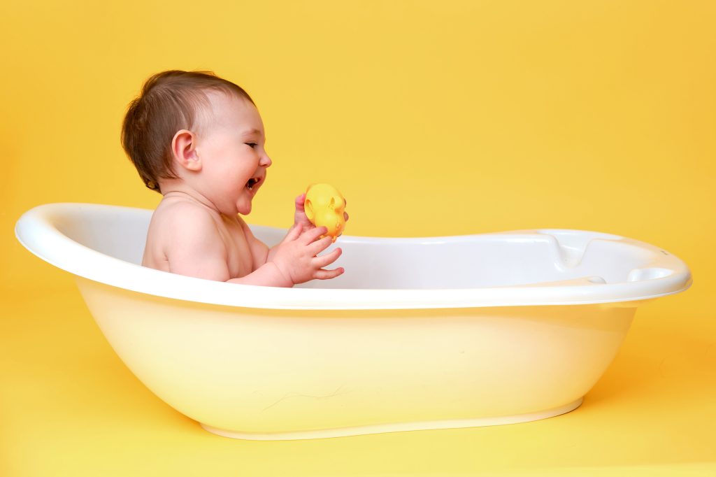 Happy baby toddler boy playing with a yellow duck toy in a white bathtub on a studio yellow background. A smiling child at the age of one year, copy space