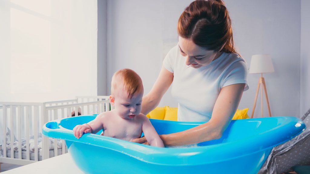 happy mother supporting infant son taking bath in baby bathtub