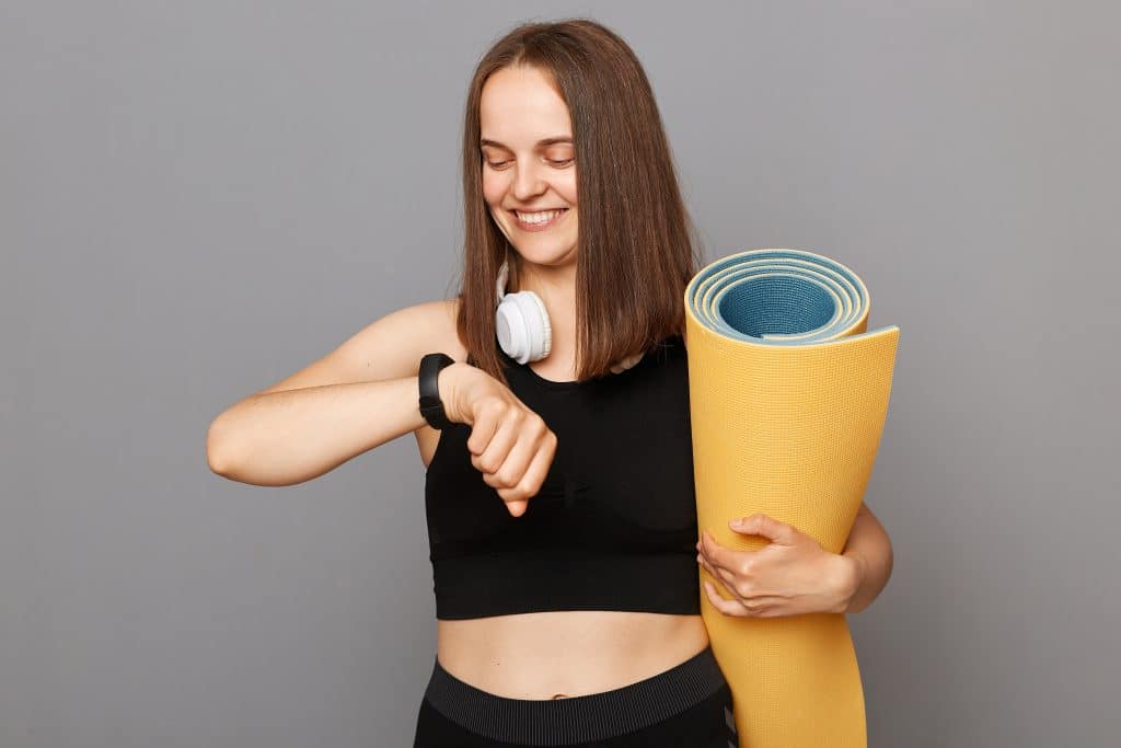 Smiling young girl wearing black top holding fitness mat in her hands posing isolated over gray background checking burned calories on her fitness tracker.