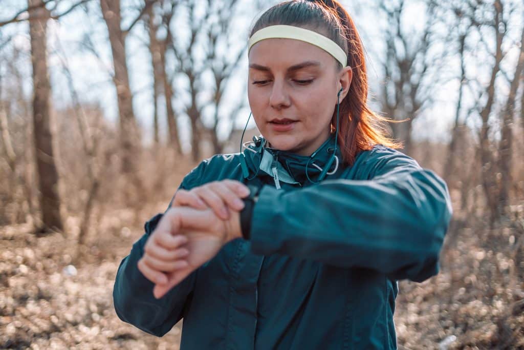 Sport watch run woman checking smartwatch tracker. Trail running runner girl looking at heart rate monitor smart watch in forest wearing jacket sportswear. Female athlete jogger training in forest