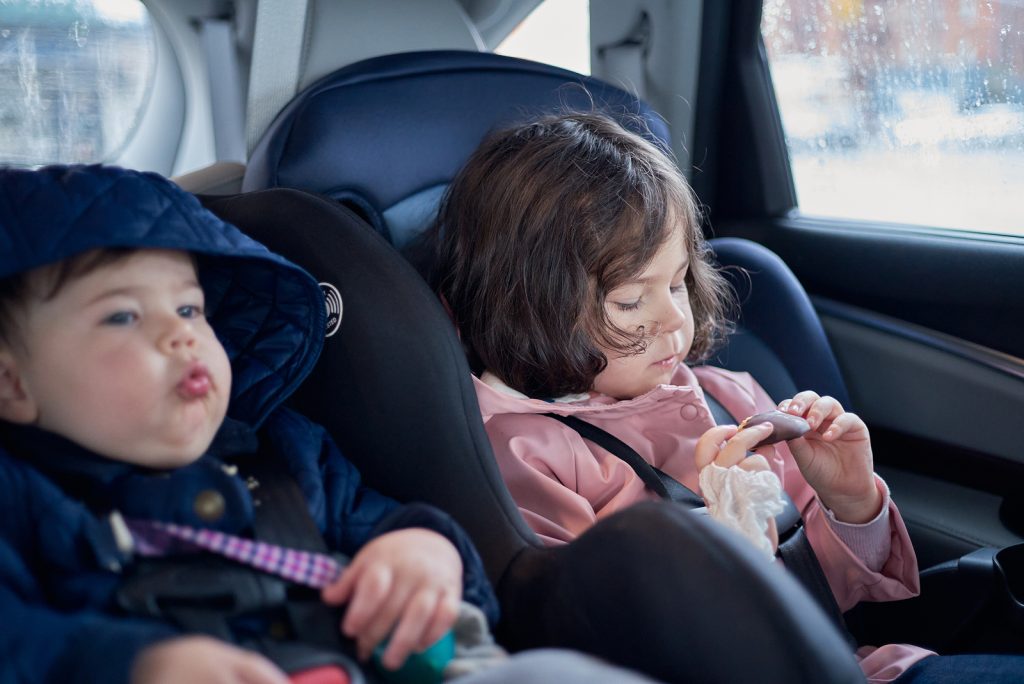siblings in car seats having snacks in the back seat of a car on a rainy day